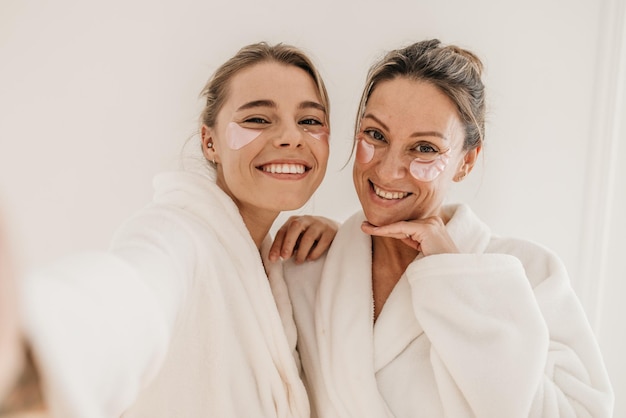 Free photo happy caucasian women use eye patches, wear bathrobes and look at camera on white background. concept of preserving youth