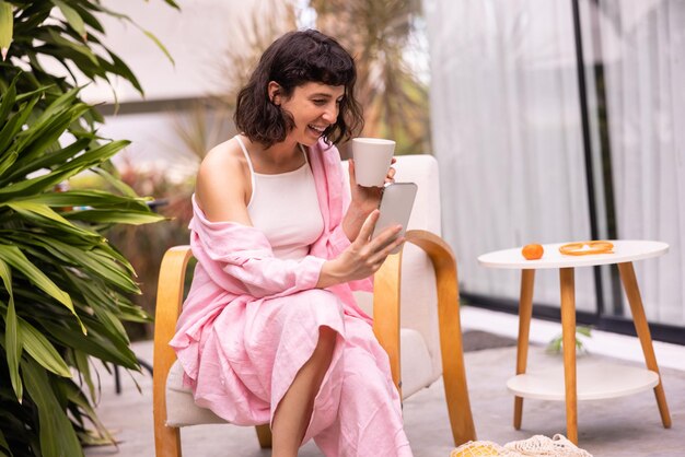 Happy caucasian young brunette woman looking at phone screen and enjoying morning coffee Summer lifestyle concept