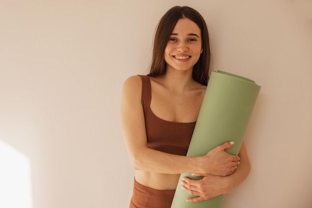 Free photo happy caucasian young girl holding yoga mat smiling at camera on beige background pretty woman trainer in sports tight clothes healthy lifestyle concept