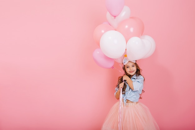 Happy celebration of birthday party with flying balloons of charming cute little girl in tulle skirt smiling to camera isolated on pink background. Charming smile, expressing happiness. Place for text