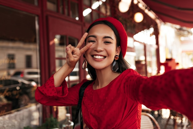 Free Photo happy charming brunette woman in red dress, stylish beret and eyeglasses smiles sincerely, shows peace sign and takes selfie outside