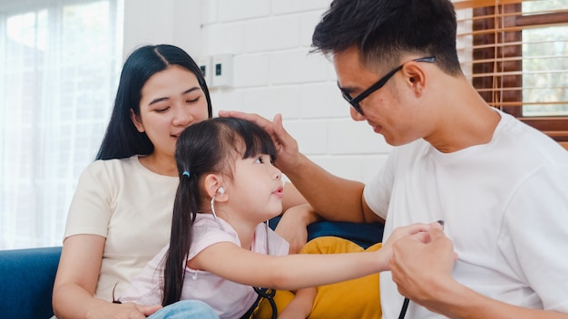 Happy cheerful asian family dad, mom and daughter playing funny game as doctor having fun on sofa at home