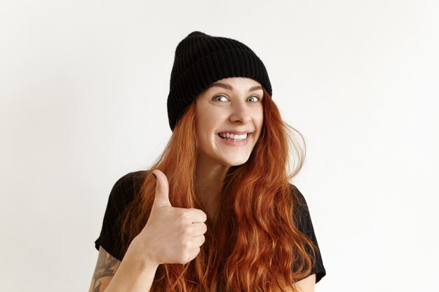 Happy cheerful European girl with tattoo and messy hairstyle, showing thumbs-up gesture