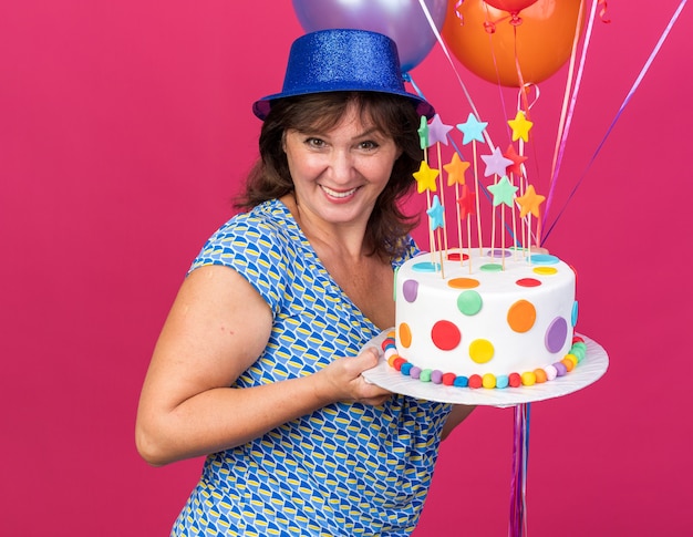 Happy and cheerful middle age woman in party hat with colorful balloons holding birthday cake  smiling broadly