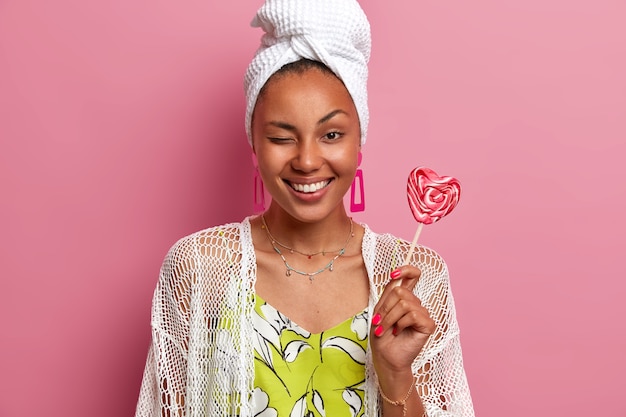 Happy cheerful woman feels refreshed after taking shower, wears wrapped towel on head, has healthy skin, white teeth, winks eye and smiles broadly, holds lollipop, isolated on pink wall