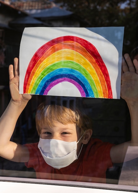 Free Photo happy child with medical mask holding rainbow sign through the window at home