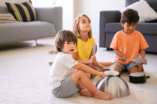 Happy children sitting on carpet and playing with utensils. Cute Caucasian little boys and blonde girl having fun together in living room and knocking on pans. Childhood and home activity concept