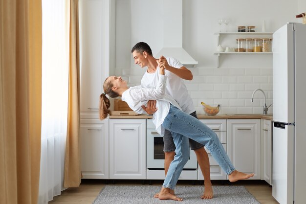 Happy couple dancing in the kitchen, husband and wife wearing white shirts dance in morning near the window, expressing love and romantic feelings, indoor shot.