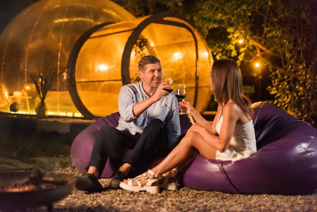 Free photo a happy couple drinking wine near a fireplace at night transparent bubble tent on the background