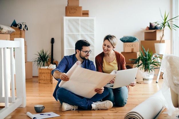 Happy couple examining blueprints and using laptop at their new apartment