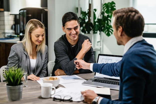 Happy couple having a meeting with real estate agent and analyzing blueprints while communicating