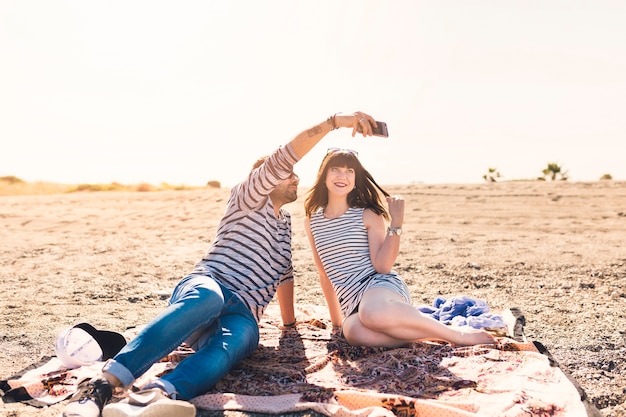 Happy couple sitting on beach taking self portrait on mobile phone