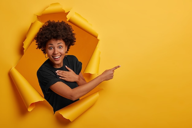 Happy curly haired African American woman laughs positively, points aside on copy space, wears black t shirt