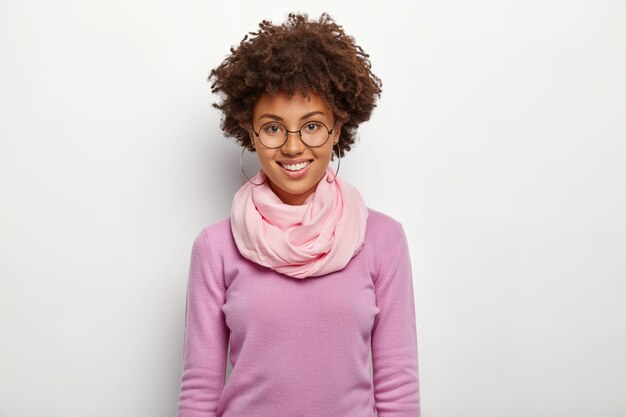 Happy curly woman wears spectacles and violet clothes, enjoys awesome day, smiles gently