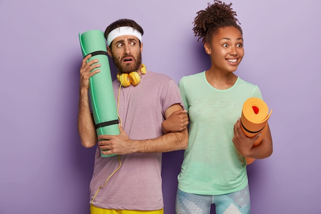 Free photo happy curly young afro american woman looks positively aside, holds hand of boyfriend, carries mat, puzzled worried man wears headband, uses headphones connected to some device