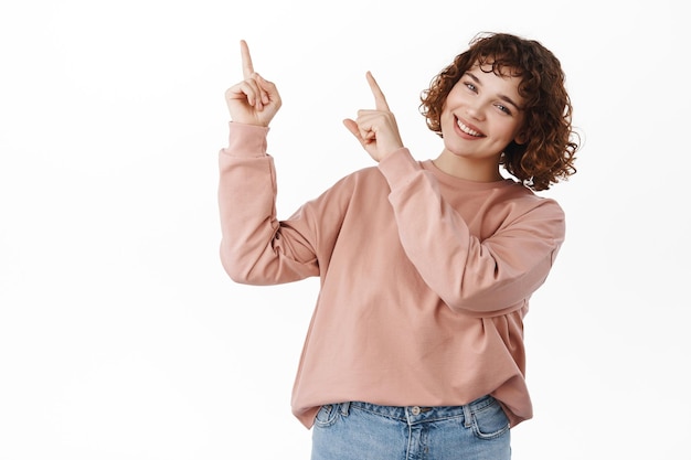 Happy cute girl pointing at upper left corner banner, showing logo with announcement or discount info, smiling broadly at camera, standing against white background.