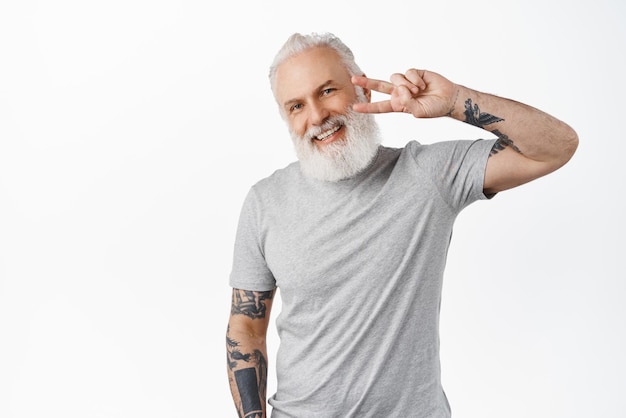 Happy cute senior guy with tattoos and beard shows peace vsign looking kawaii and smiling at camera standing in grey casual tshirt against white background