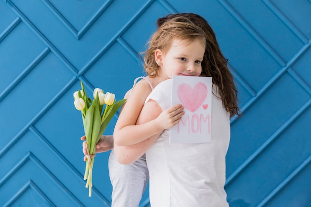 Free photo happy daughter hugging her mother with holding flowers and greeting card against blue backdrop