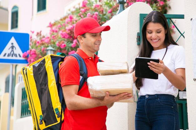 Happy deliveryman standing near customer with tablet. Professional postman in red uniform holding boxes and delivering order. Pretty female client getting parcels. Delivery service and post concept