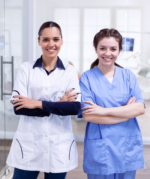 Happy dentistiry coworkers smiling standing in line Portrait of stomatology team in dental reception with arms crossed looking at camera wearing uniform.