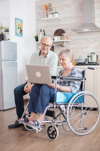 Happy disabled woman during video conference in the kitchen. Disabled senior woman in wheelchair and her husband having a video conference on tablet pc in kitchen. Paralyzed old woman and her husband