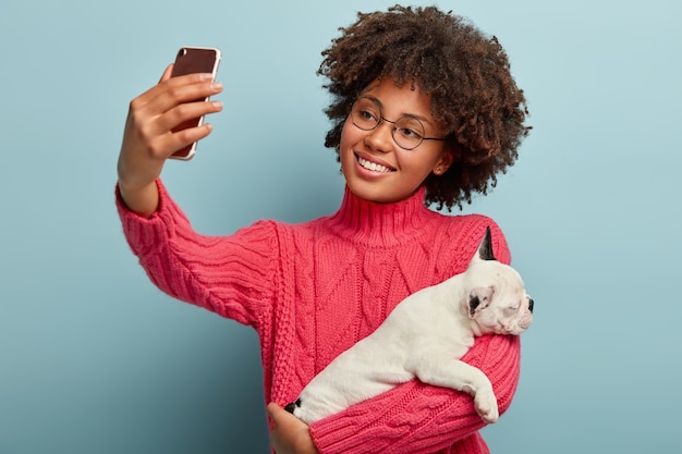 Happy dog owner tilts head, has Afro hairstyle, dressed in pink knitted jumper, holds sleeping little puppy