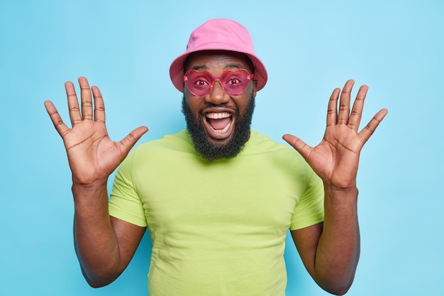 Happy emotional bearded guy raises palms feels very glad exclaims loudly reacts on amazing news wears stylish pink sunglasses casual t shirt and panama isolated over blue wall