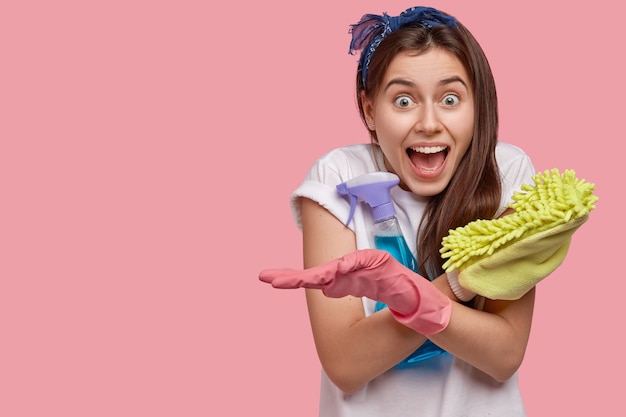 Happy emotional woman crosses hands, holds mop and spray cleanser, wears white t shirt and gloves, glad to finish housework in time, not late for date, poses against pink wall. Good mood for cleaning