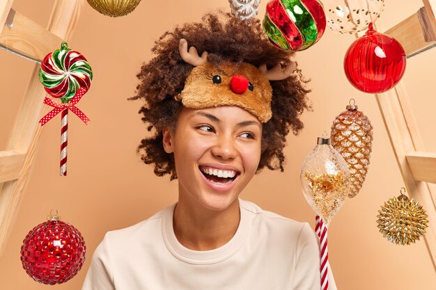 Happy emotions and festive mood. Positive sincere woman smiles broadly expresses positive emotions wears reindeer sleep mask on forehead surrounded by New year toys spends time at cozy home.