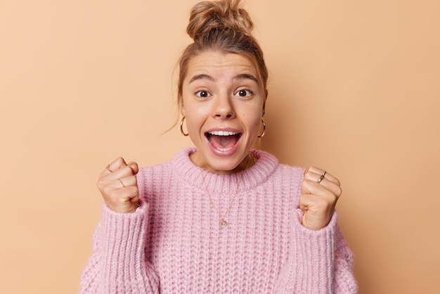 Free Photo happy energetic young woman clenches fists from joy exclaims and feels like winner celebrates achievement has combed hair wears knitted sweater isolated over beige background roots for someone