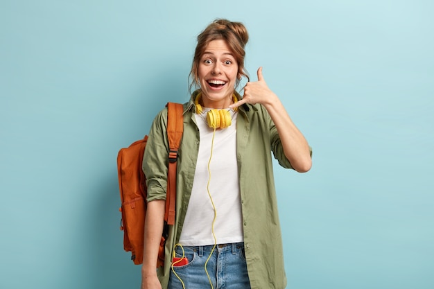 Happy European woman makes call gesture, tries to contact with friend on distance, wears modern headphones