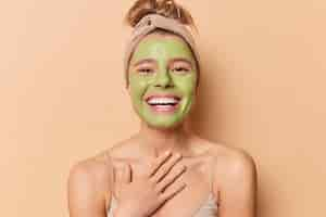 Free photo happy european woman smiles broadly keeps hand on chest applies nourishing green mask undergoes beauty treatmentss wears headband has combed hair isolated over brown background skin care concept