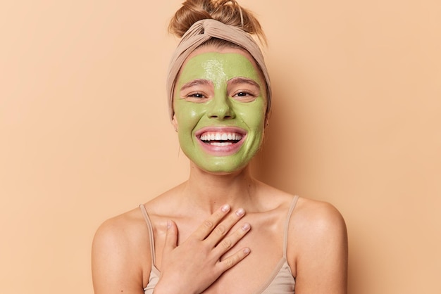 Happy European woman smiles broadly keeps hand on chest applies nourishing green mask undergoes beauty treatmentss wears headband has combed hair isolated over brown background Skin care concept