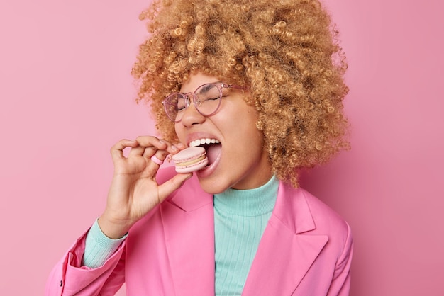 Happy European woman with curly hair bites appetizing macaroon has addiction to sugar dressed in elegant clothes wears transparent glasses poses against pink bacground Confectionary concept