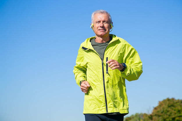 Free photo happy excited senior man in wireless headphones jogging outside. low angle, blue clear sky. front view, copy space. activity and age concept