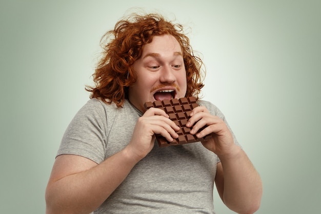 Happy excited young chubby red-haired male opening mouth widely while biting bar of chocolate, feeling impatient. Funny Caucasian man in grey t-shirt consuming unhealthy but delicious junk food