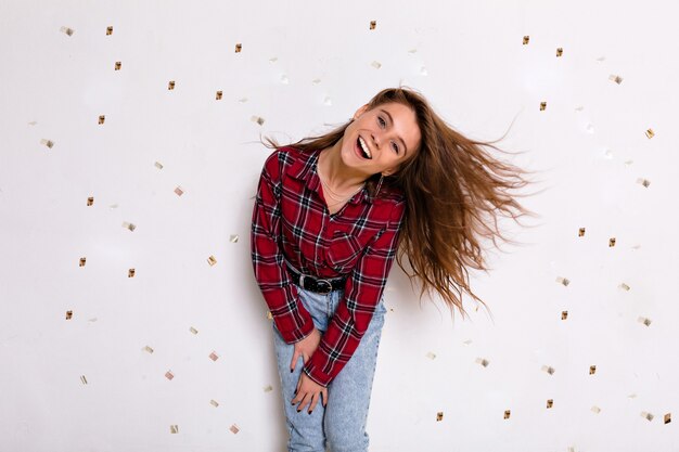 Happy exited lovely woman has fun and dancing over white wall with confetti in casual outfit. Excited lady looking at fallen paper hearts and laughing.