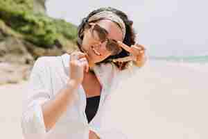 Free photo happy exited young woman with dark wavy hair wears black glasses and hair ornament in white shirt walks down the white sandy beach in sunlight