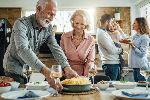 Happy extended family having lunch in dining room. Focus is on mature couple brining food at the table.