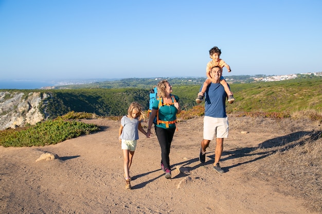 Happy family couple and children  hiking in countryside, walking on path. Excited boy riding on dads neck. Full length. Nature and recreation concept