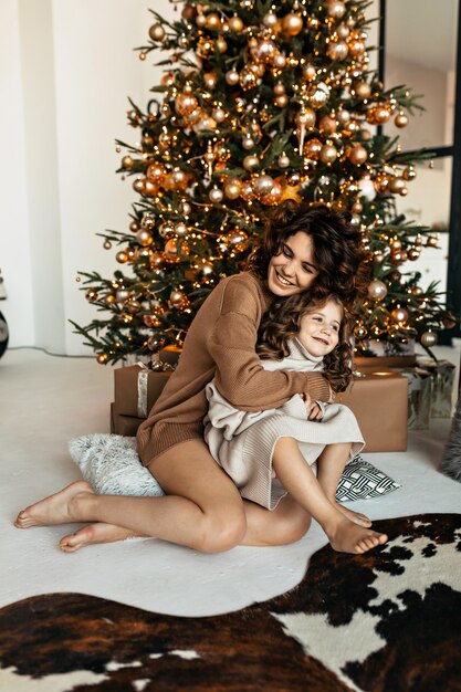 Happy family portrait of pretty mom with daughter dressed knitted sweaters sitting in front of Christmas tree and celebrating New Year