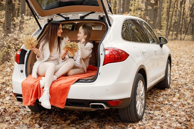 Free photo happy family resting after day spending outdoor in autumn park. mother and her child girl sitting inside white car trunk. mother and daughter wearing white clothes.