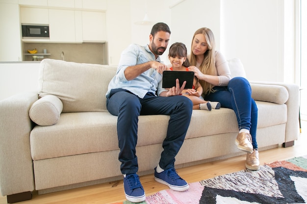 Happy family sitting on couch, using online app on tablet, looking at screen, watching movie together.