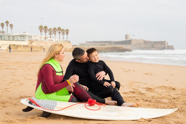 Happy family sitting on sand near surfboard