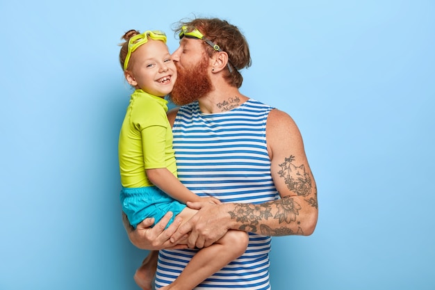 Free photo happy father and daughter wear goggles and summer clothes, have fun together during rest. affectionate dad carries litle girl, kisses her in cheek, expresses love. family and recreation concept