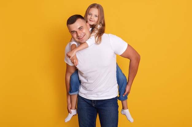 Free photo happy father and little girl wering white t shirts and jeans, posing isolated on yellow, have happy facial expression, spending time together. family concept.