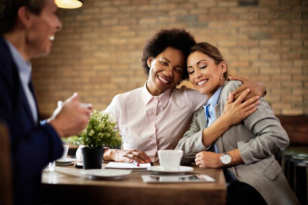 Happy female colleagues embracing with eyes closed while having coffee break in a cafe