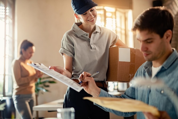 Happy female courier holding clipboard while businessman is signing for a delivery in the office