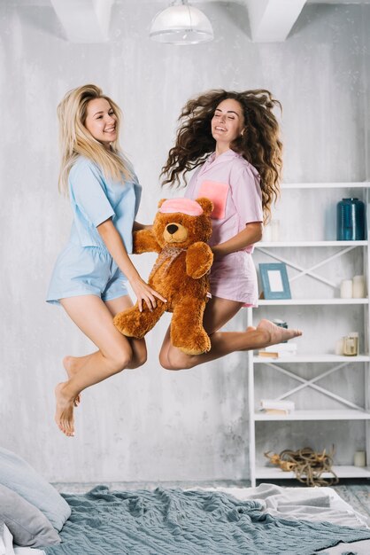 Happy female friends jumping over bed with soft toy