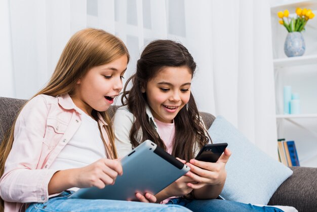 Happy female friends sitting on sofa using digital tablet and mobile phone